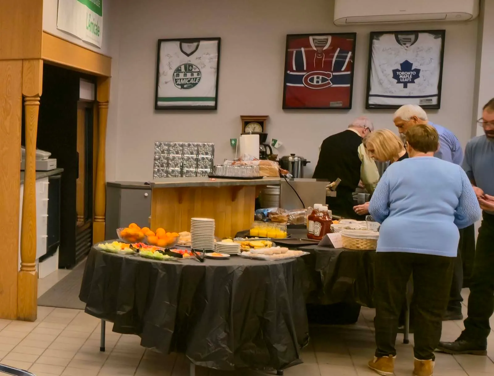 Table de service du repas à L’Amicale, avec des membres se servant nourriture et boissons sous des chandails sportifs encadrés au mur.