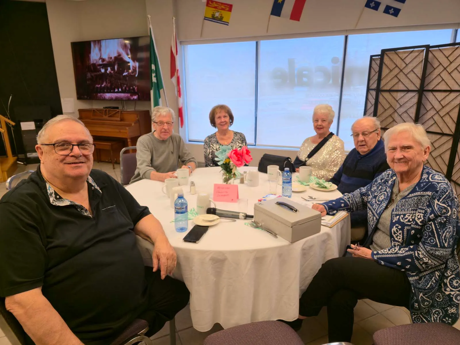 Groupe de membres de L’Amicale assis autour d’une table ronde, souriant à la caméra pendant un moment de partage et de discussion.