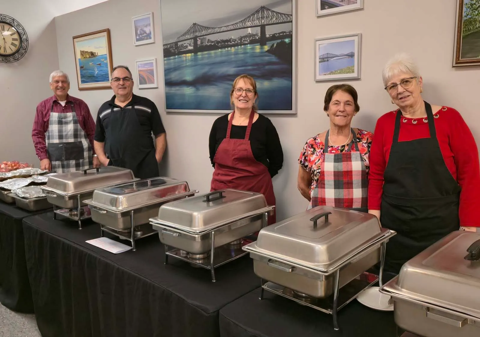 Groupe de membres bénévoles de L’Amicale debout derrière le buffet, souriant devant les plats chauds prêts à être servis.