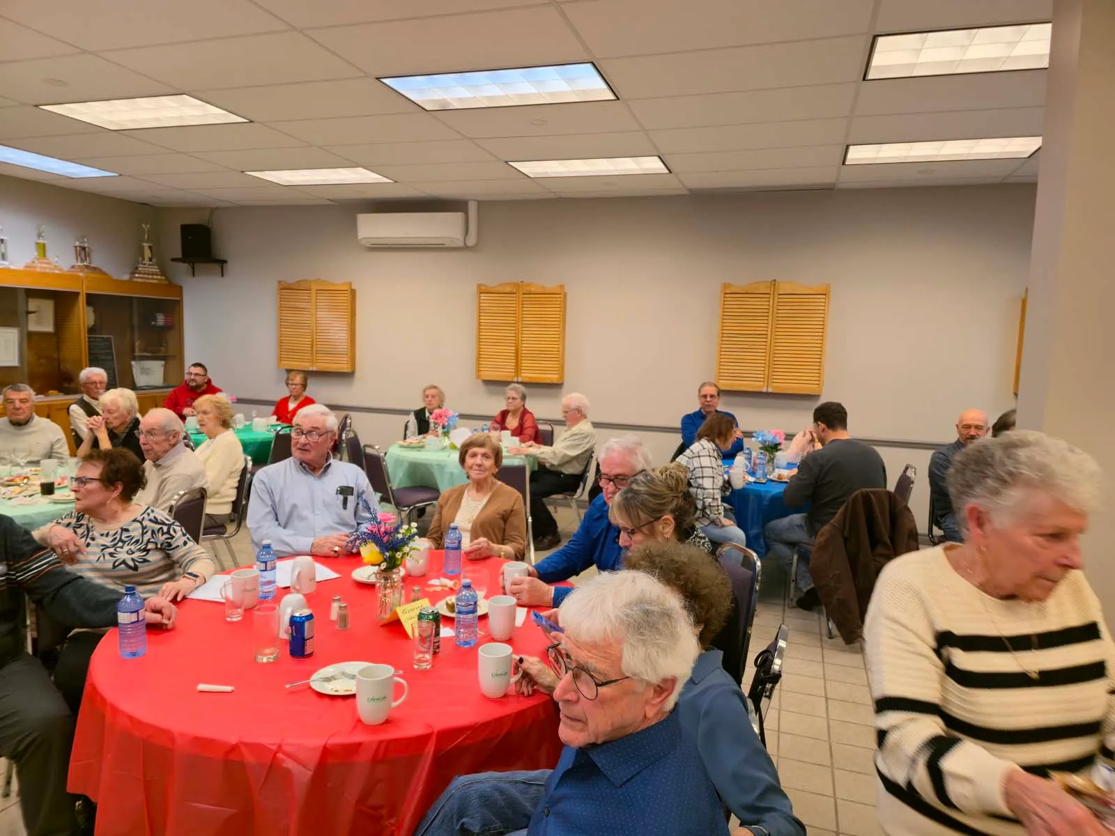 Membres de L’Amicale assis autour de tables colorées, écoutant une animation dans une ambiance chaleureuse.