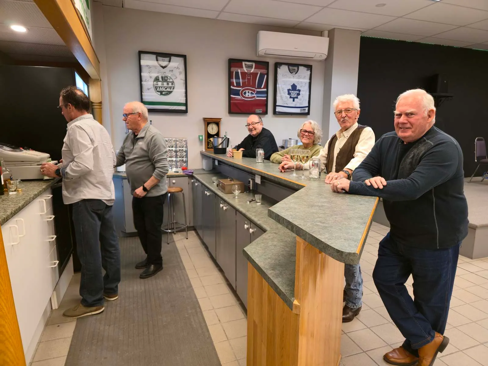 Membres de L’Amicale rassemblés autour du comptoir, discutant et partageant un moment convivial avant le repas.