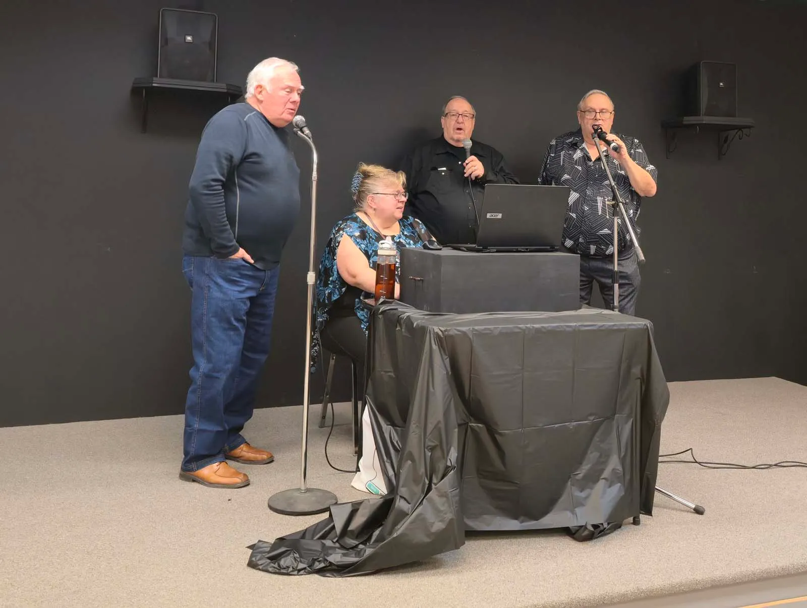 Groupe de membres de L’Amicale chantant sur scène, accompagnés d’un ordinateur et de micros lors d’une animation musicale.