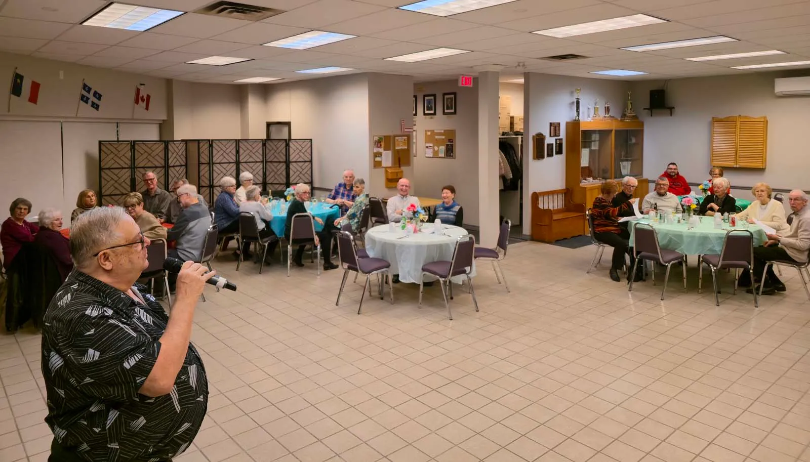 Un membre de L’Amicale tenant un micro, s’adressant aux membres assis autour des tables dans la salle.