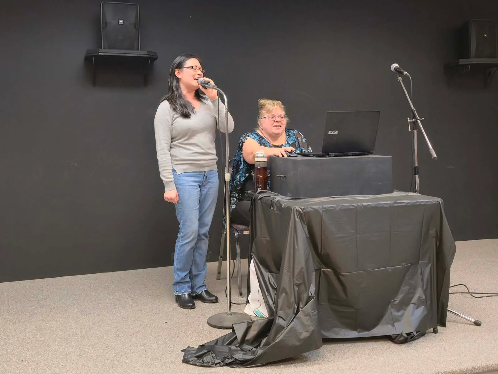 Deux membres de L’Amicale chantant au micro, accompagnés d’une musicienne assise devant un ordinateur sur scène.