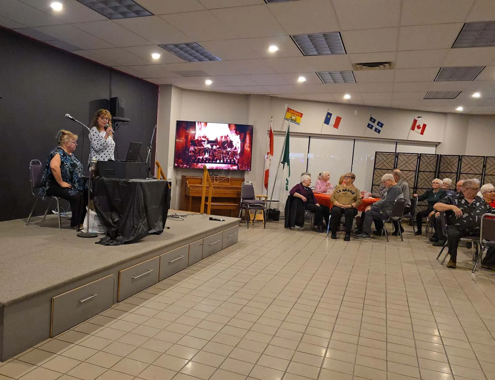 Vue d’ensemble de la salle de L’Amicale avec des membres assis et attentifs pendant une animation musicale.