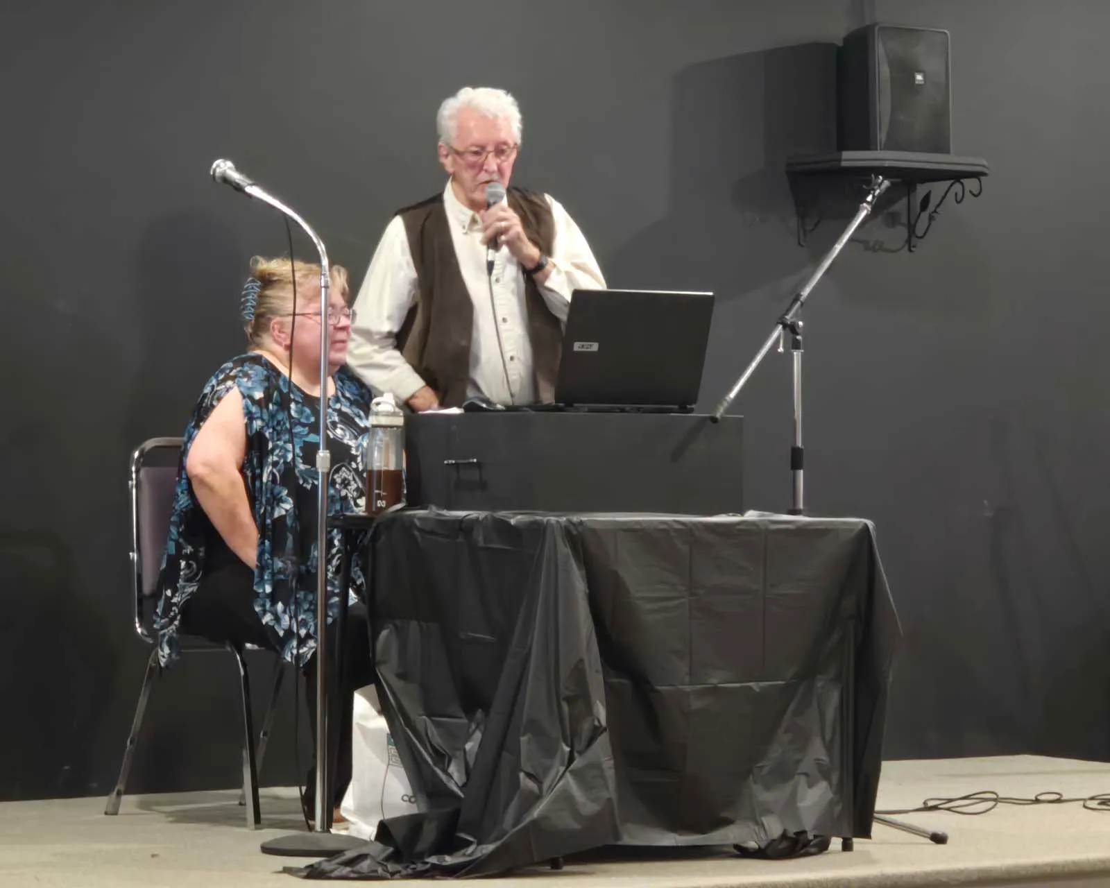 Membre de L’Amicale chantant au micro sur scène, accompagné d’un poste audio et d’une animatrice assise.