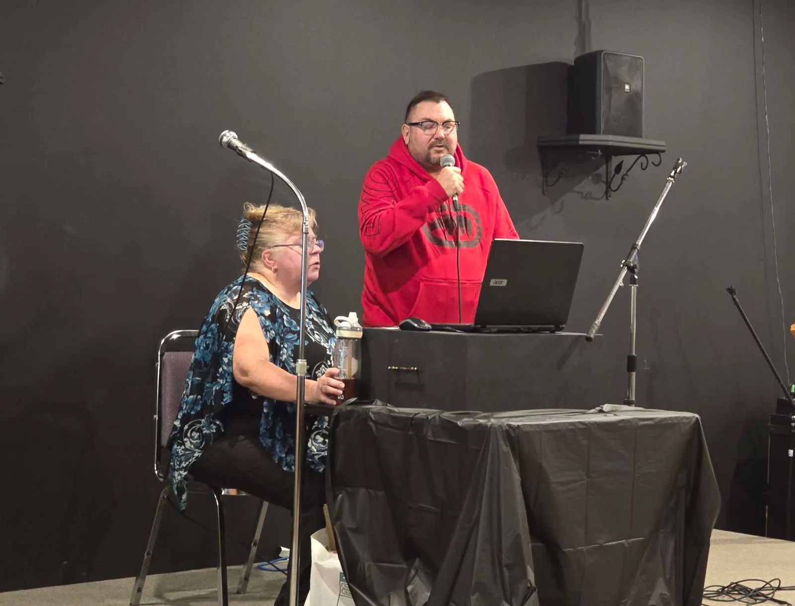 Membre de L’Amicale chantant au micro sur scène, accompagné d’une animatrice au poste audio.