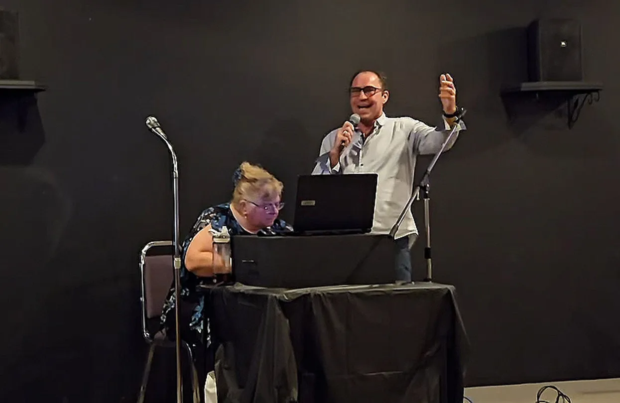 Un membre de L’Amicale chante sur scène pendant que l’animation musicale est gérée à l’ordinateur.