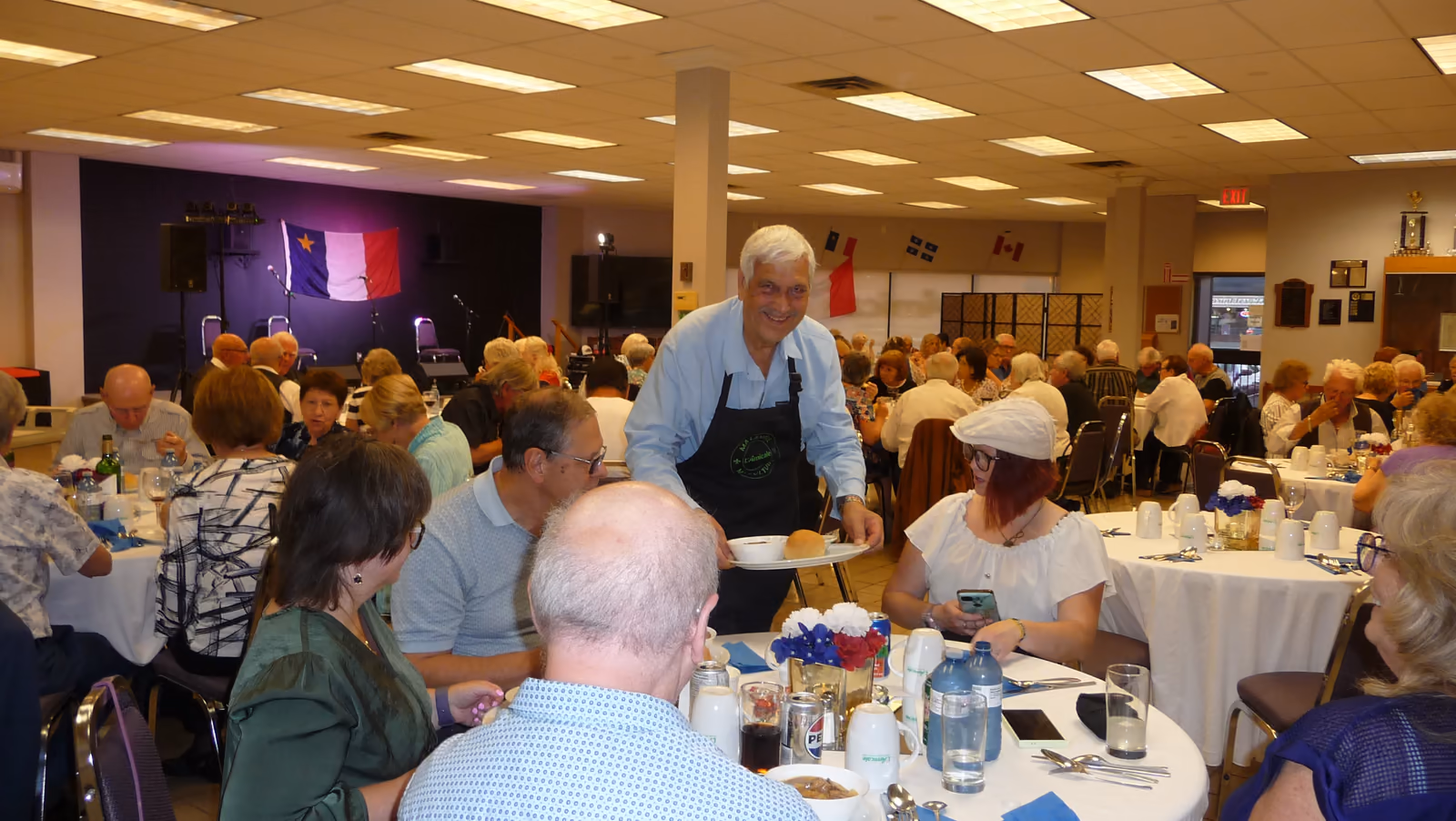 Un bénévole en tablier sert le souper acadien aux convives attablés dans la grande salle.