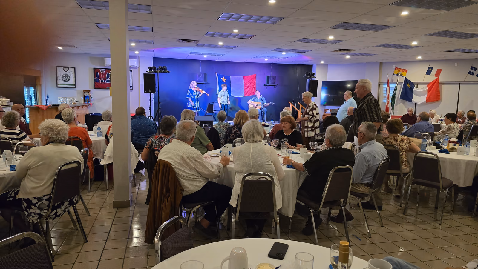 Le public attablé assiste au spectacle de la famille Goguen, sur scène sous le drapeau acadien.