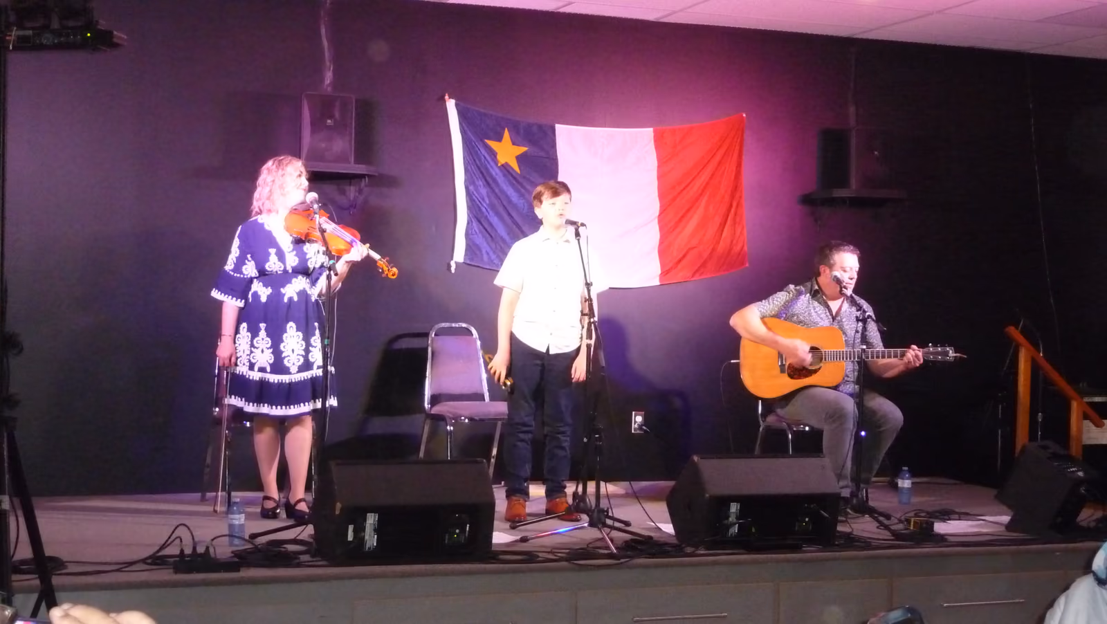 Une violoniste, un jeune chanteur et un guitariste de la famille Goguen jouent sur scène devant le drapeau acadien.