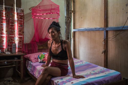 A woman wearing a black bikini top and a reddish skirt or shorts sits on a bed’s multicolored blanket