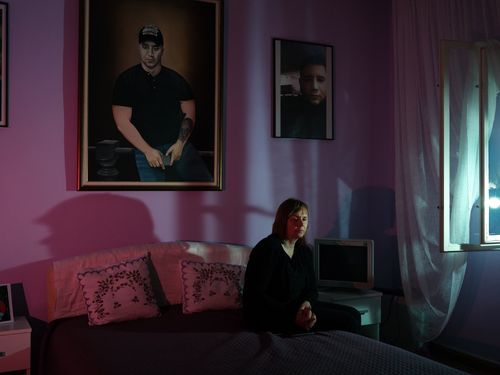 A woman wearing a black top sits in front of a purple wall where a giant portrait of a young man hangs.