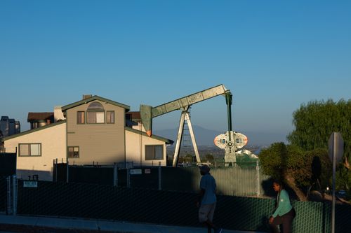 Two people pass a building and an oil derrick.