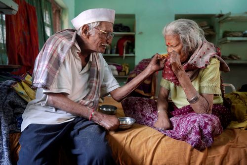 An elderly man feeds his wife, who is sitting cross-legged next to him. He feeds his wife by hand; both are missing fingers due to the effects of leprosy.