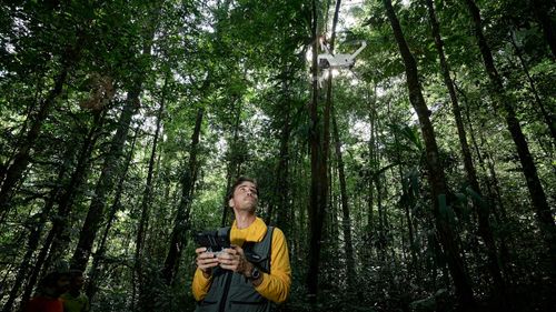 A man stands in a forest holding a drone controller. The drone is visible overhead, with a green background of tall trees.