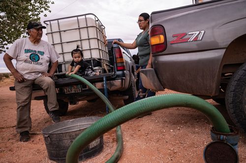 A man filling an underground tank with water sits on the back of a truck with a baby. A young woman stands at right.