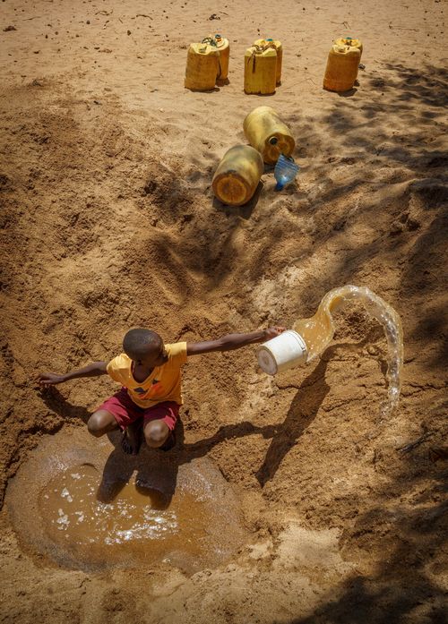 A young person uses a white bucket near dirty water.