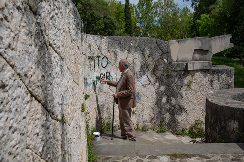 An older man with a cane and brown suit stands in front of a stone wall.