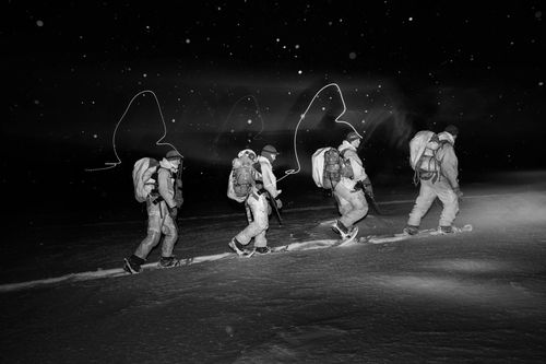 Four people in winter gear hike through a snow-covered landscape at night. They carry backpacks and use headlamps, which create trails of light.
