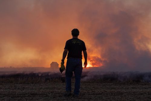 A person stands in a field, facing a large wildfire with intense smoke and flames in the distance. The sky is filled with a smoky haze.