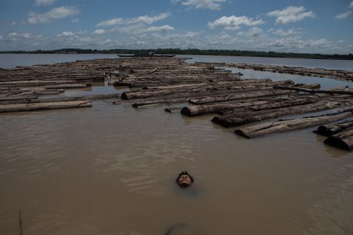 A man bathes in a river next to logs, with just his face above the water, under a partly cloudy sky.