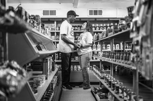 A black-and-white image of a man and woman standing in a grocery store aisle, smiling at each other. Both wear matching shirts with a logo. Shelves are filled with various canned and bottled products.