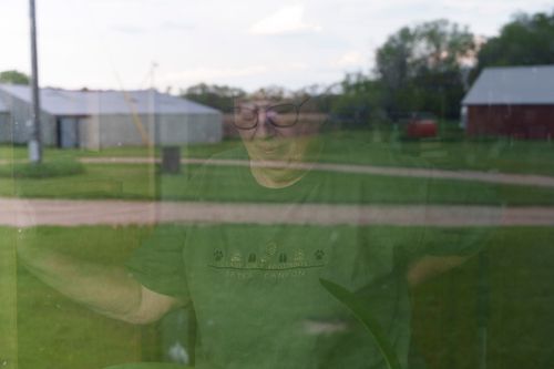 A person wearing glasses is seen through a reflective window, standing in an outdoor setting with buildings and trees in the background.