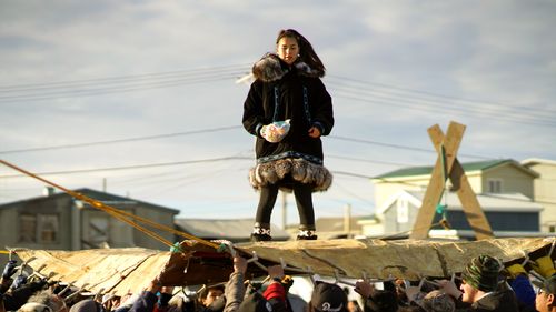 A person in a thick coat with fur stands on a large seal skin being held up by a group of people. The background shows a few houses and a partly cloudy sky.