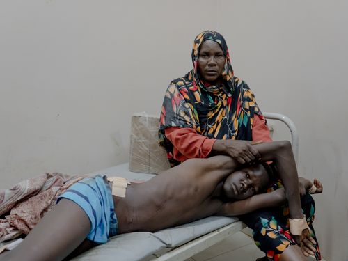 A woman wearing a colorful patterned head scarf sits on a hospital bed, cradling a shirtless boy with bandaged wrists. The boy lies partially on her lap. The room is bare.