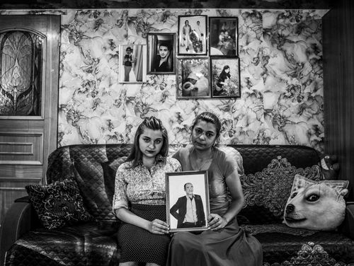 Two women sit on a floral-patterned sofa, holding a framed photo of a man. Behind them, a wall is decorated with family portraits. A pillow shaped like a dog's face rests on the couch.