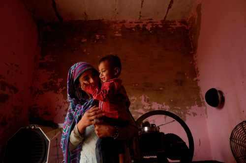 A woman wearing a colorful headscarf stands holding a smiling child. The child is wearing red.