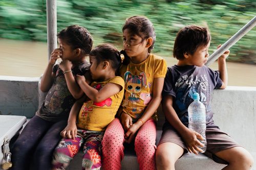 four small children sit on a boat, with a river and the rainforest blurred in the background.