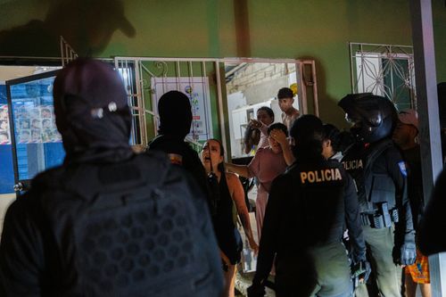 A group of masked police officers confront a woman outside a house. The woman appears to be shouting, while others, including a man and a child, observe from the doorway.