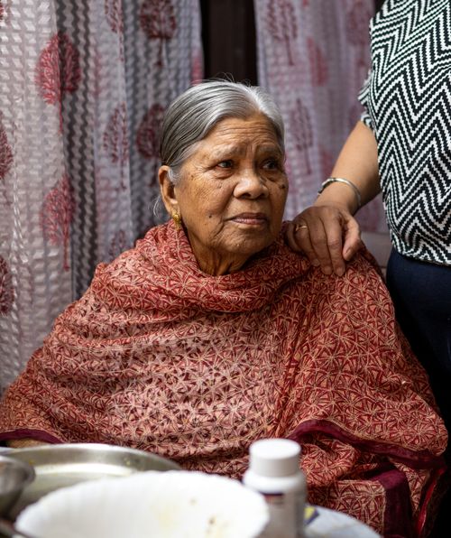 An elderly woman with gray hair sits at a table with a hand on her shoulder, wearing a brown shawl.