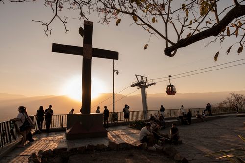 A large cross stands on a hilltop at sunset, surrounded by people looking at the view. A cable car travels in the background. The sky is a warm gradient of orange and yellow.