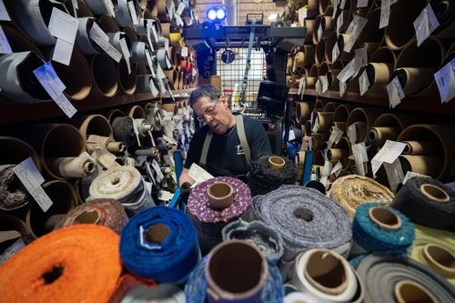 A man stands surrounded by shelves filled with rolls of various fabrics. He appears to be taking inventory or inspecting the materials, with the colorful rolls prominently displayed in the foreground.