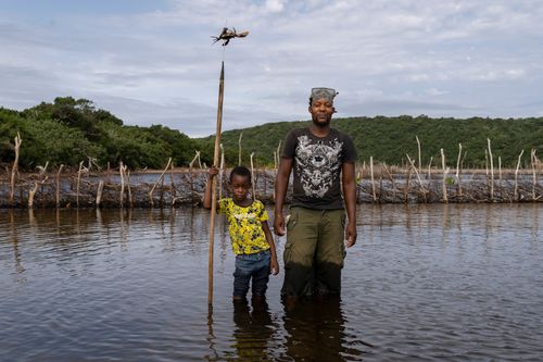 A man and his young son stand knee-deep in water, the boy holding a spear for spear fishing.