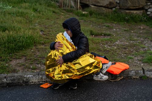 A person in a black hooded jacket sits on a roadside, holding a child wrapped in a gold emergency blanket. An orange life vest and a pair of shoes lie nearby on the wet pavement.