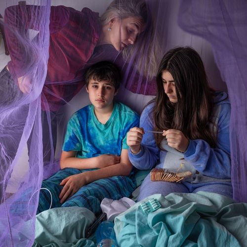 a mother looks down at her two teenage children in bed. One is looking at a thermometer and has crackers and pills in her lap.