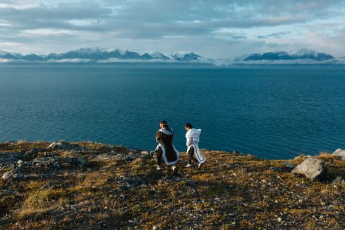 Two people in long coats walk along a grassy cliff overlooking a large body of water. Snow-capped mountains and a cloudy sky are visible in the background.