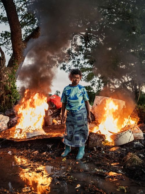 a woman stands in front of a pile of burning trash, with the fire reflected in puddles on the ground.