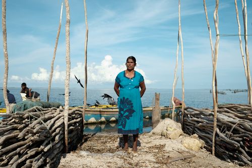 A fisherwoman stands on the beach in front of a boat. She is wearing a blue dress, and holds her hands behind her back.