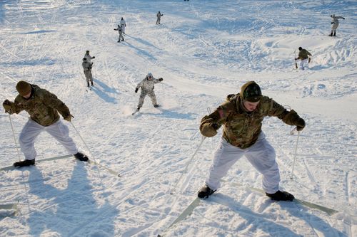 A group of eight soldiers in camouflage coats and white pants ski uphill in the snow.