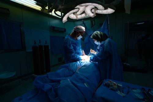 Two doctors perform surgery on a patient in a dimly lit operating room illuminated by overhead lights. Both surgeons are wearing blue scrubs and protective masks.