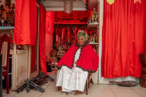 a woman sits for a portrait in her home, dressed in red-and-white Afro Brazilian clothing, with figurines and red curtains in the background.