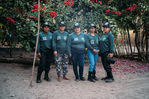 Five women in matching green uniforms and caps stand in a row outdoors. They are standing in front of red flowers and greenery.