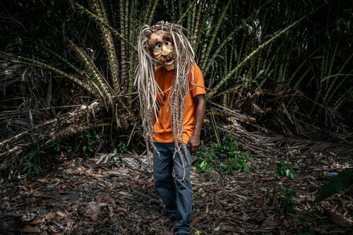 A person stands in front of dense foliage wearing a mask with long hair-like strands and a mustache.