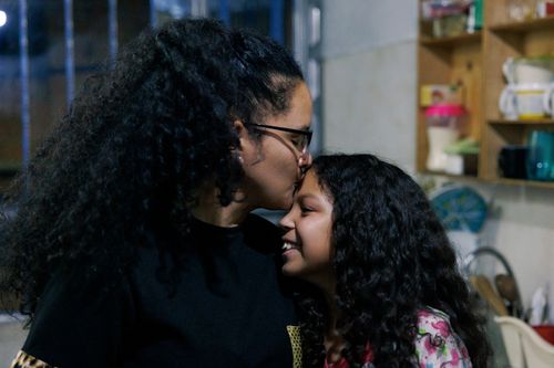 A woman with long curly hair kisses a young girl with similar hair on the forehead. They are standing in a kitchen with shelves holding cups and containers in the background. The girl is smiling.
