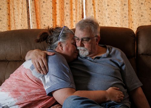 An elderly couple sits closely on a couch, embracing each other, with closed eyes and relaxed expressions.